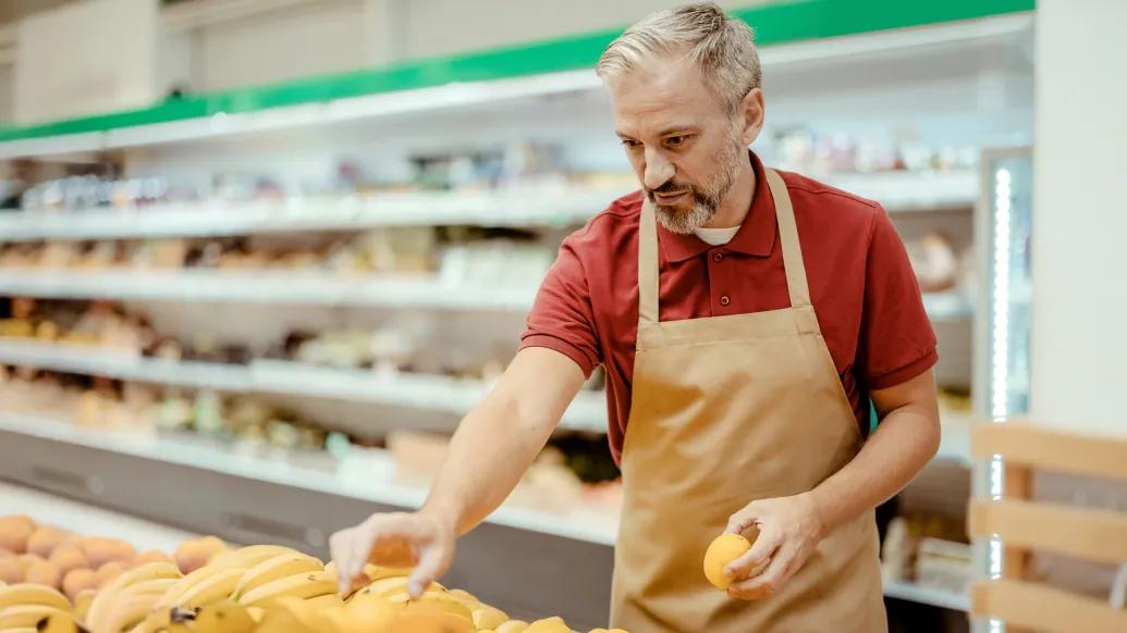 man at grocery store packing produce shelves red shirt beige apron
