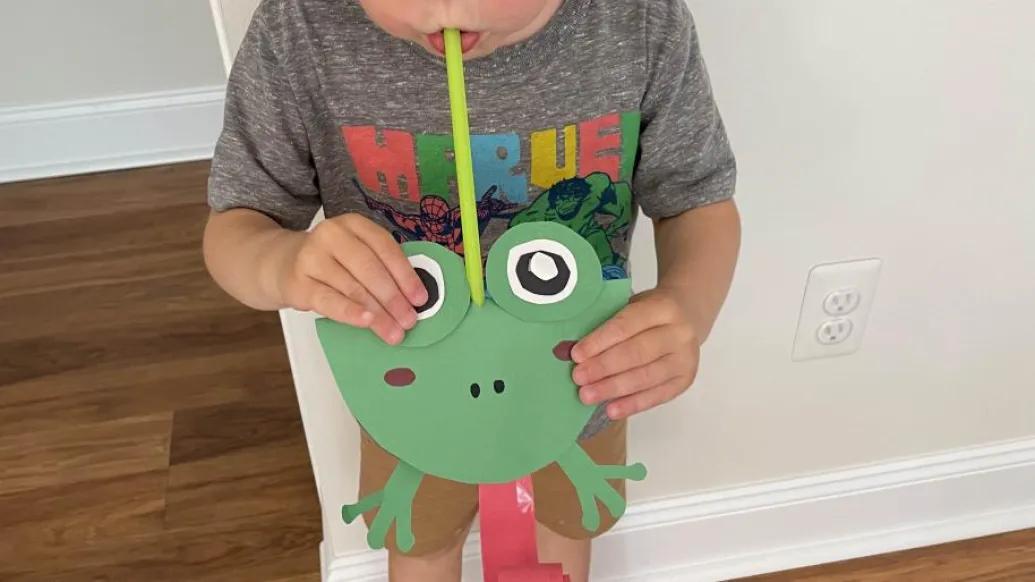 child blows straw on frog-shaped paper craft