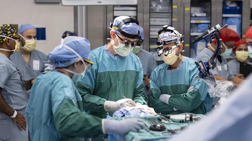 A team of medical professionals in surgical attire performs a procedure in an operating room. They are surrounded by medical equipment, including a robotic arm and various monitors.