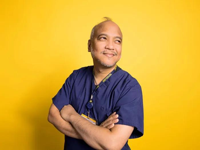 Brown-skinned man wearing dark blue scrubs with arms crossed standing against maize background