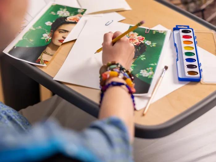 Woman's arm with colorful bracelets holding a pencil with art prints and paintbox on table
