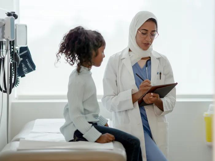 Female doctor wearing glasses and white hajib talking to young girl in exam room