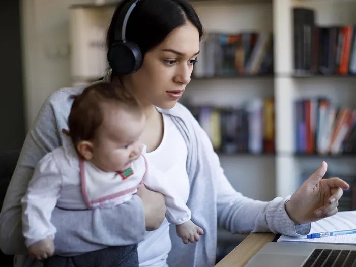 Young mother with dark hair wearing headphones, holding a baby, and looking at a laptop