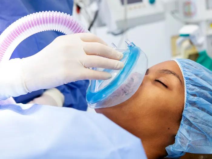 An unconscious woman lies on a hospital table while a clinician's glove is seen placing an oxygen mask over her nose and mouth