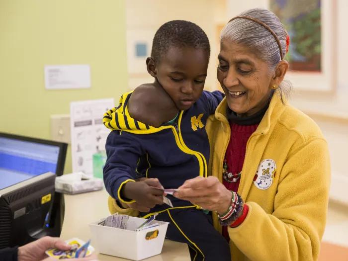 Little boy Jonathon from Uganda wearing blue M jacket, being given sticker by brown-skinned woman in maize jacket