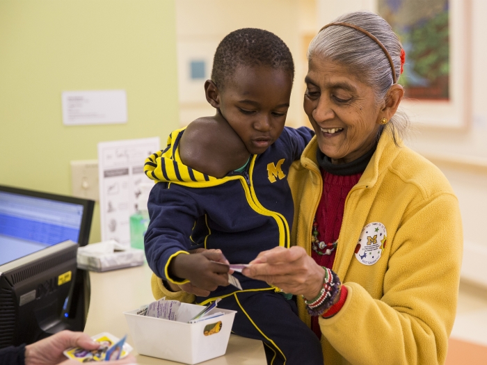 Little boy Jonathon from Uganda wearing blue M jacket, being given sticker by brown-skinned woman in maize jacket