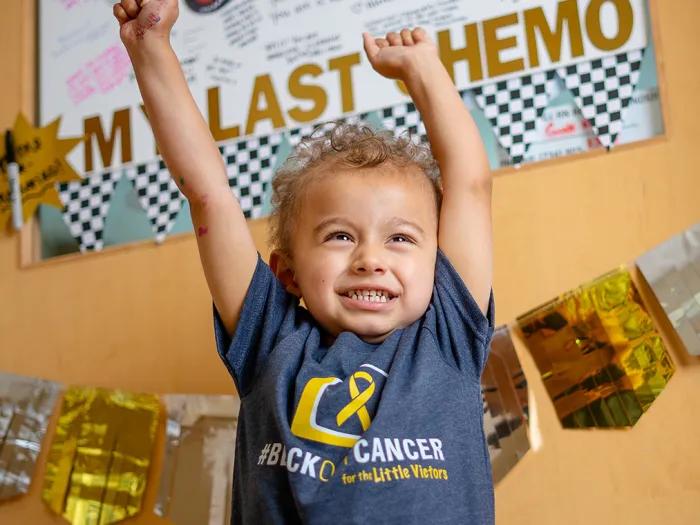 Little boy showing excitement with raised arms over his head. He is wearing a block out cancer t-shirt.