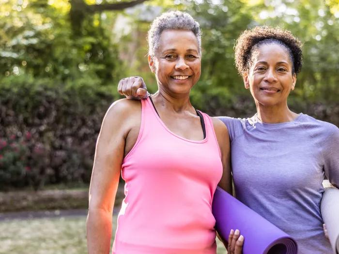 two Black women standing next to eachother, one in a pink tank top, one in long sleeve purple shirt, with yoga purple matts