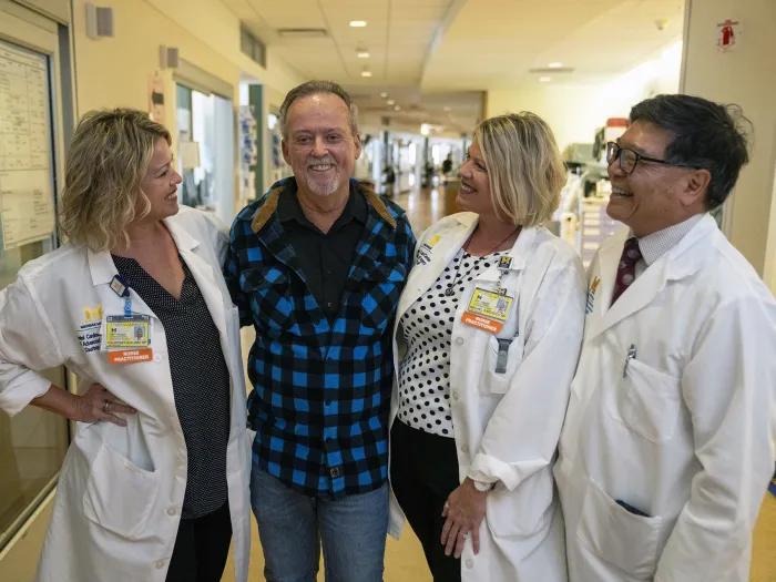 A smiling patient stands in a hospital hallway with three members of his care team in white coats. The group is standing close together, smiling warmly, showing a supportive relationship.