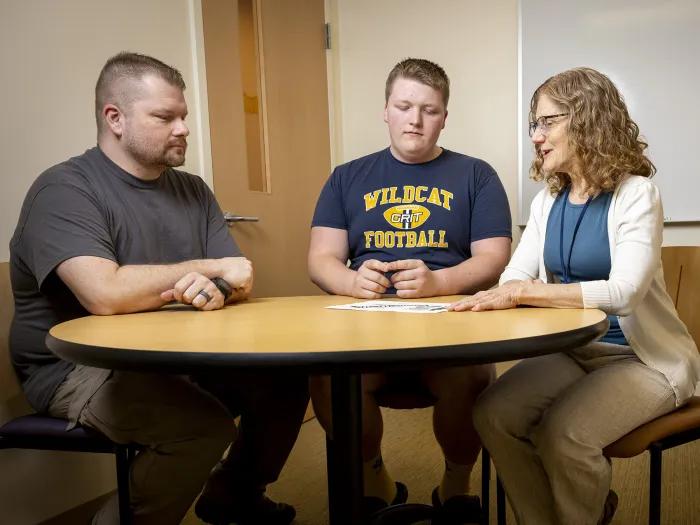 A teenage boy and his father sit at a round table with a healthcare provider, who is explaining information on a paper. The group looks engaged in discussion inside a clinic room.