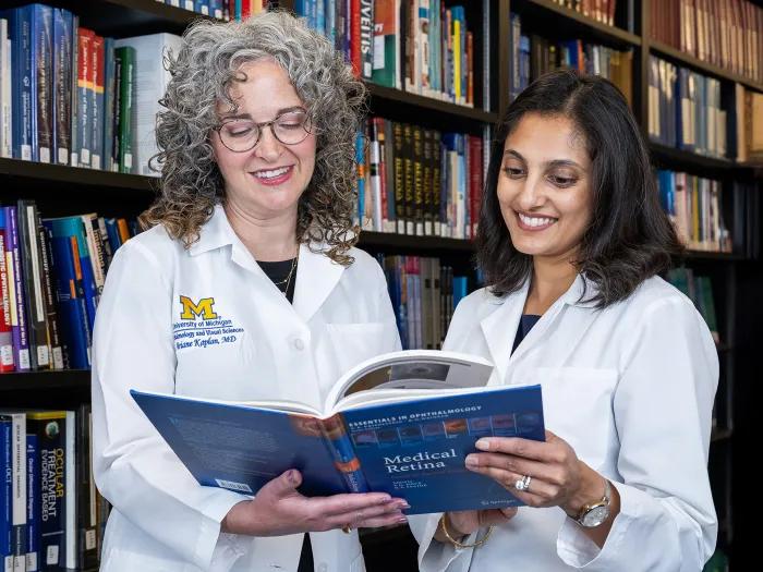 Ariane Dev Kaplan, MD and Anjali Shah, MD looking at a book and standing in front of shelf full of books