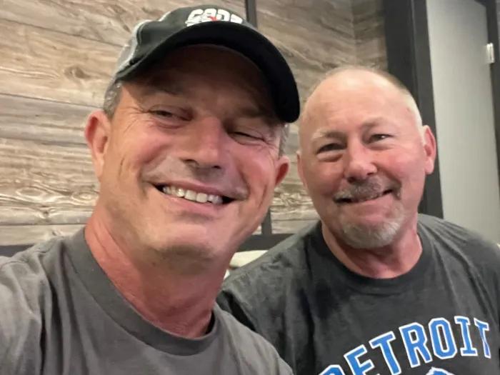 Two men smiling into camera, one wearing baseball cap the other without hat wearing Detroit Lions tshirt