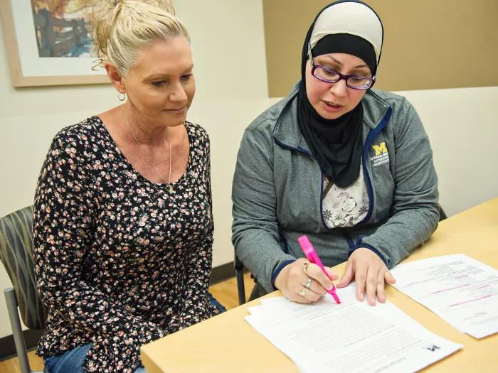 Two women seated at table reviewing a form, one with a pink highlighter and wearing a head covering and glasses