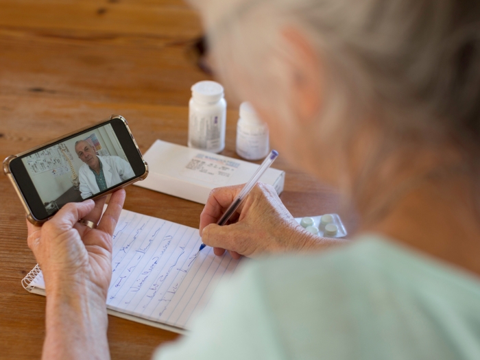 An older woman seen from behind holds a phone on which a doctor is talking to her while she takes notes with pill bottles nearby 