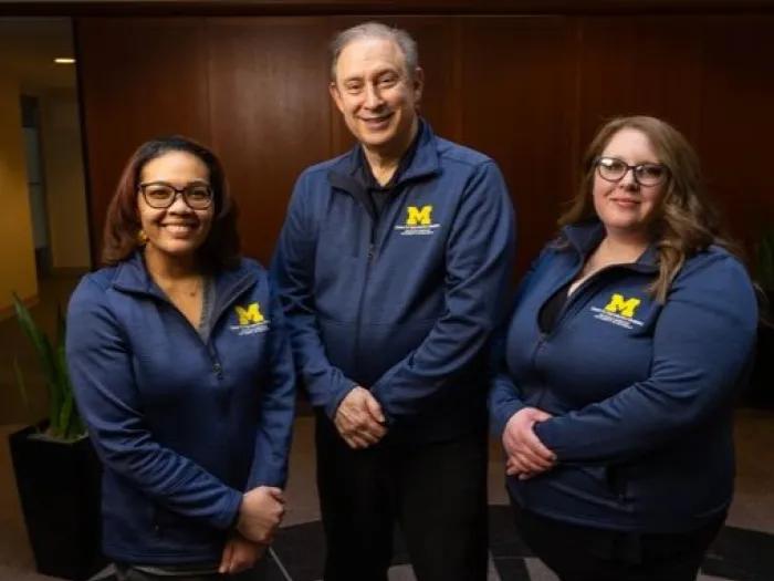 Three healthcare staff members wearing navy University of Michigan jackets stand together and smile at the camera in an indoor setting with wood-paneled walls.