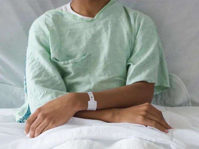 The torso and arms of a thin brown-skinned adolescent male in a hospital gown and bed