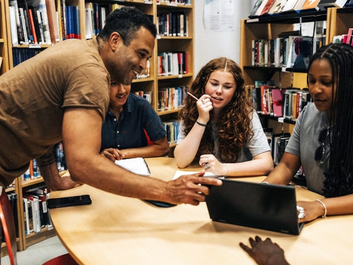 teacher with students at table looking at laptop in library 