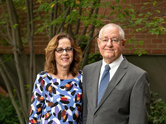 Susan Lane, wearing glasses and a dress with blue, red, black, and purple brushstroke designs, stands next to Alan Sugar, who is wearing glasses and a grey blazer with a blue tie. They are standing in a shaded courtyard area with shrubs and a brick wall behind them. 