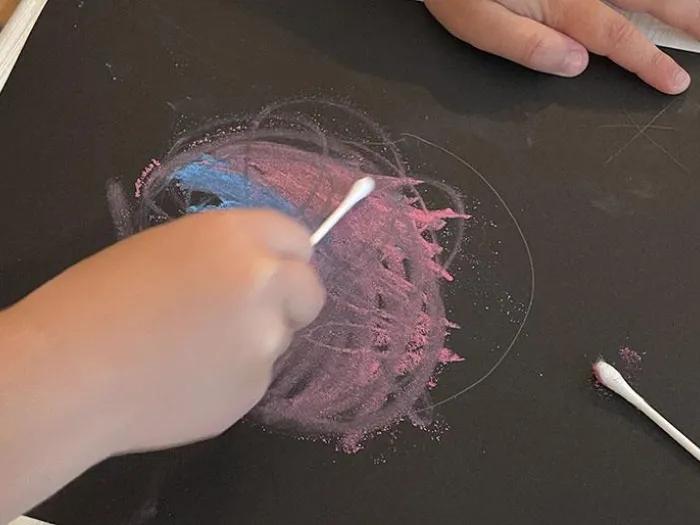 A child’s hand holding an earbud, gently smoothing a finished chalk artwork on black paper.