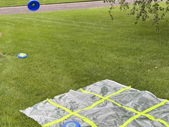 Child in yard tossing blue frisbee on plastic tic-tac-toe board divided by yellow tape