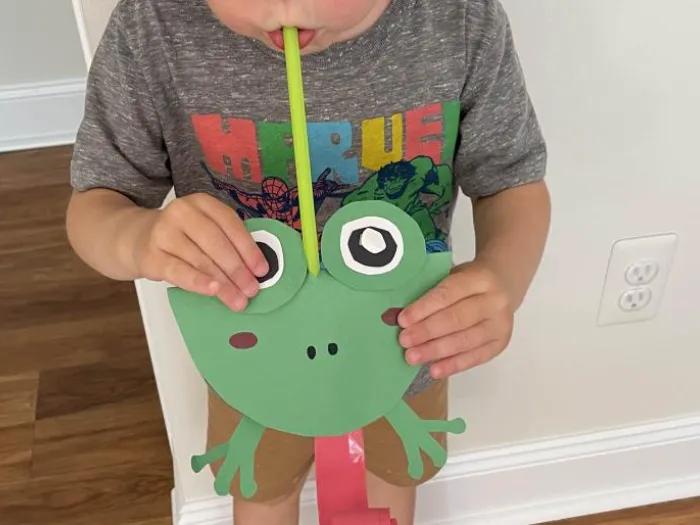 child blows straw on frog-shaped paper craft