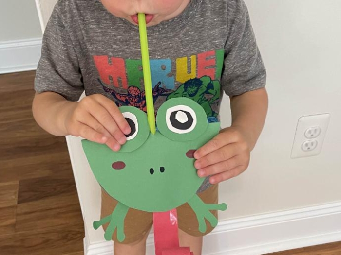 child blows straw on frog-shaped paper craft