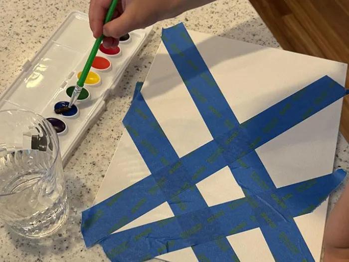 A child’s hand holds a paintbrush over a watercolor palette while painting on a white canvas taped off with blue painter’s tape in a crisscross pattern. A clear glass of water sits nearby on a speckled countertop, and part of the child’s arm and leg are visible beside the canvas.