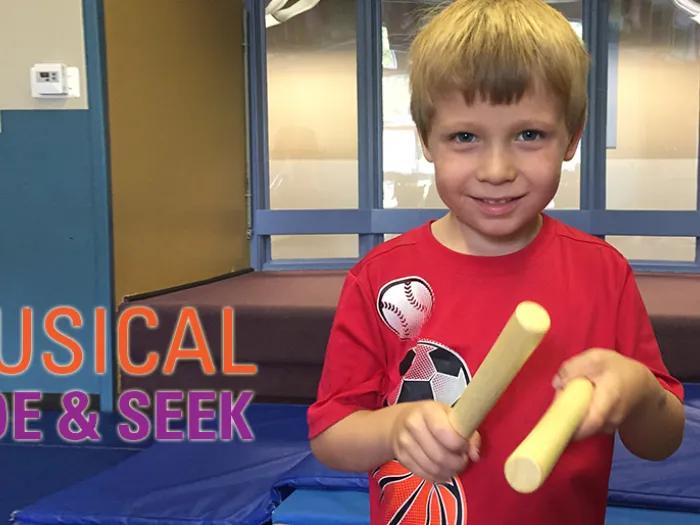 A child in red T-shirt holding two wooden sticks while playing the game of musical hide and seek.