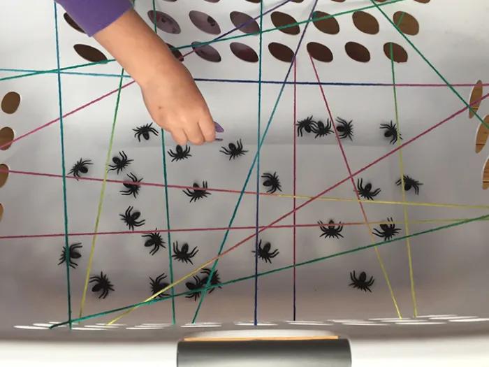 Colorful yarn attached to the holes within a white laundry basket. Child picking up plastic spiders between yarn.
