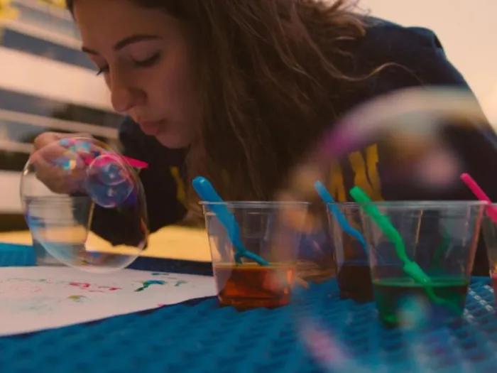 A young girl carefully painting a delicate soap bubble placed on a table, focusing intently with a fine paintbrush in her hand. Transparent glasses filled with vibrant liquid colors are arranged around the bubble, alongside scattered paintbrushes and art supplies.