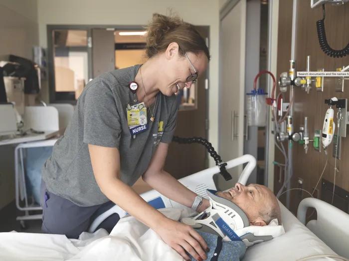 A nurse leans over the bed of a patient who is covered up and wearing a neck brace. They appear to be having a pleasant conversation.