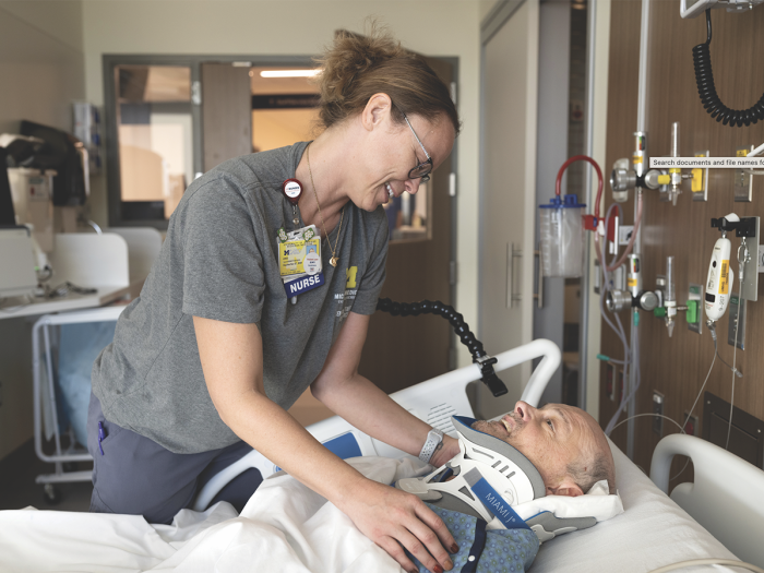A nurse leans over the bed of a patient who is covered up and wearing a neck brace. They appear to be having a pleasant conversation.