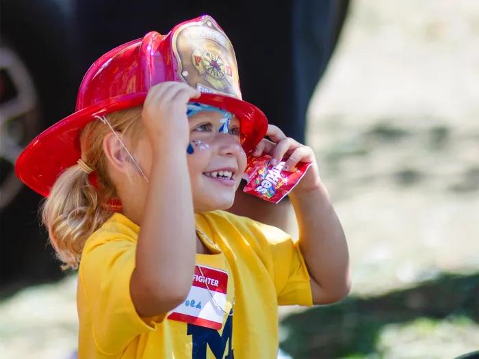 Little girl in yellow shirt with block M wearing red plastic fire hat with pack of Skittles in her hand