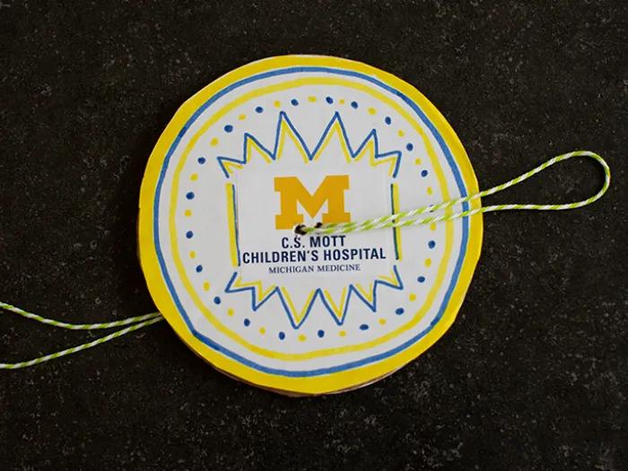 A handmade paper spinner craft featuring alternating yellow and blue sections mounted on a white rounded paper background. The center of the spinner displays a large yellow block "M" alongside the text "C.S. Mott Children's Hospital, Michigan Medicine."