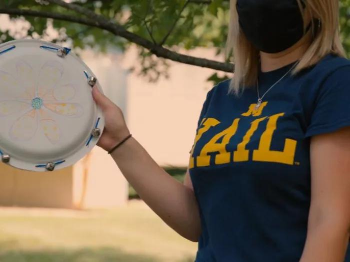 A girl wearing a blue T-shirt holds a DIY tambourine made from a paper plate.