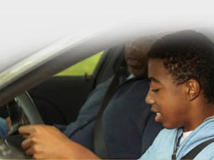 Teen driver sitting in the driver's seat with hands on the steering wheel while the father in the front passenger seat fastens his seat belt.