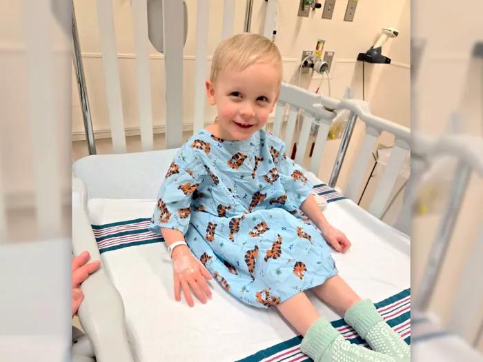 Young boy in hospital bed smiling, wearing blue hospital gown with tigers all over it and green hospital socks