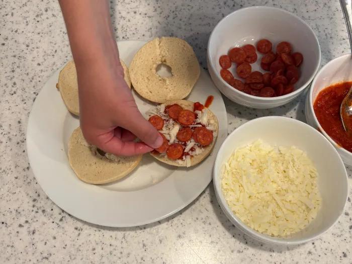 boy placing mini pepperonis on top of cheese on bagel