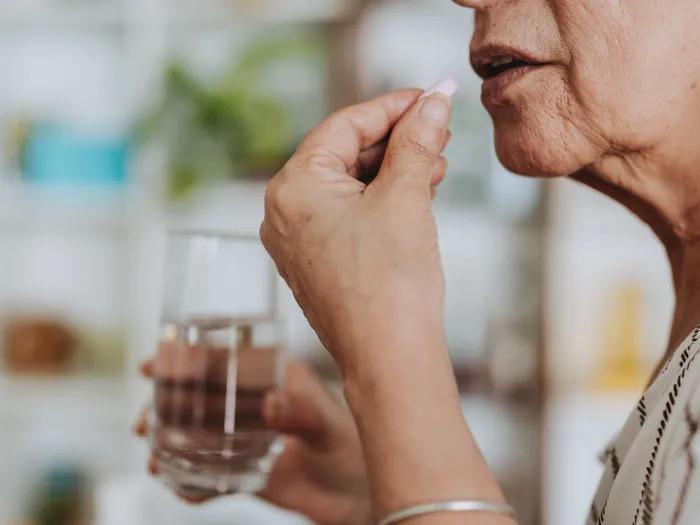 A person holds a pill near their mouth and a glass of water in the other hand, preparing to take the medication. The background is a softly focused room with shelves and blurred greenery.