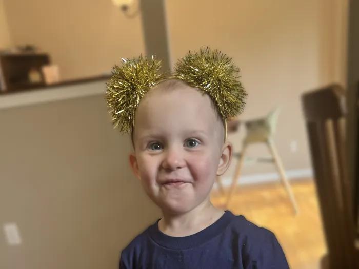 Headshot of Abel, a smiling toddler wearing a headband with two gold pompoms.
