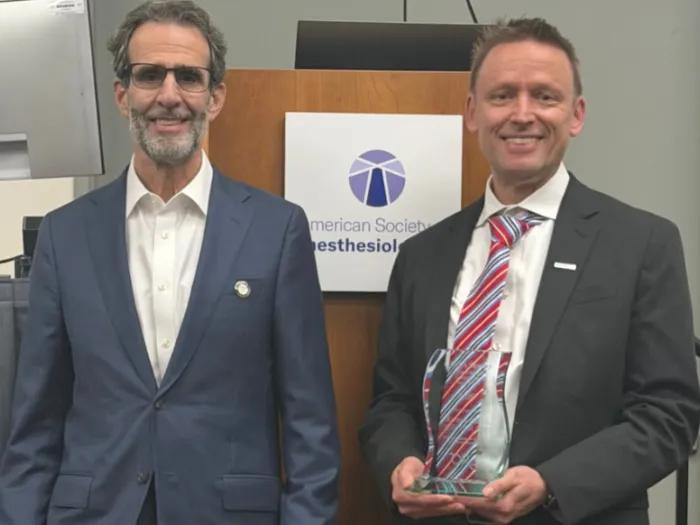 Two men in suits stand beside an American Society of Anesthesiologists sign; the man on the right holds a clear glass award.
