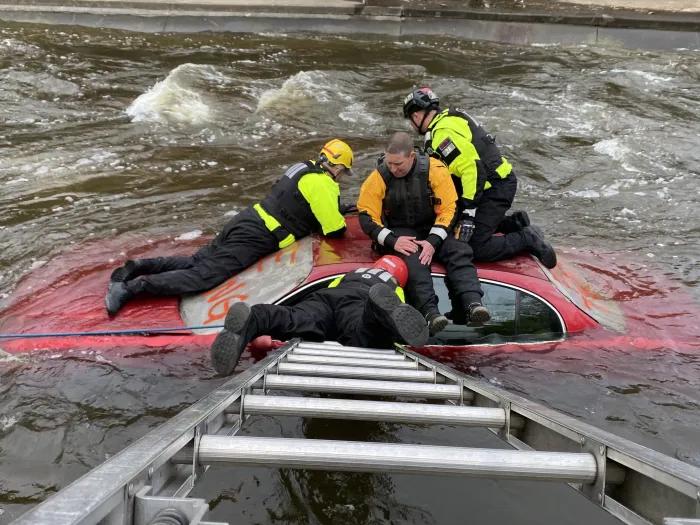 a ladder stretches to a car submerged in water. Four people sit on top the car.
