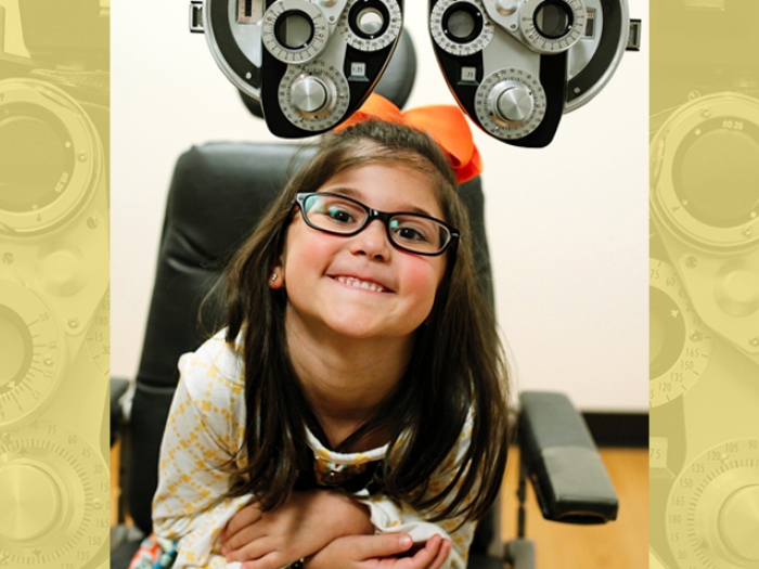 A young girl with glasses smiles while sitting in an eye exam chair under a phoropter.