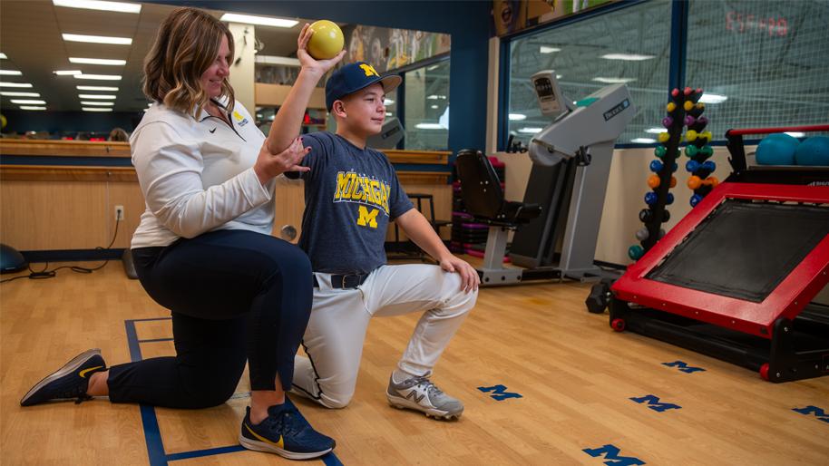 Female trainer assisting boy wearing Michigan shirt and cap to throw a yellow ball