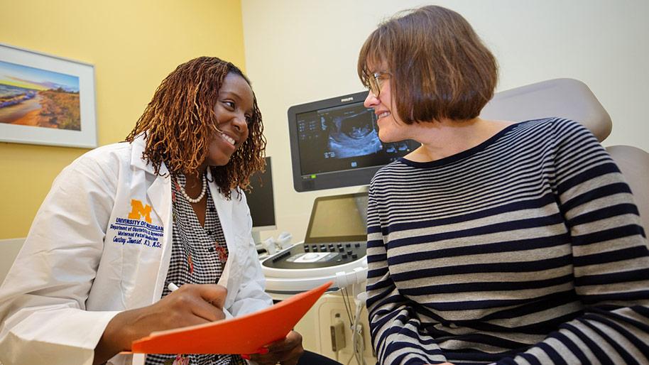 Female doctor talking to a patient in an exam room. An ultrasound image is on a monitor in the background.