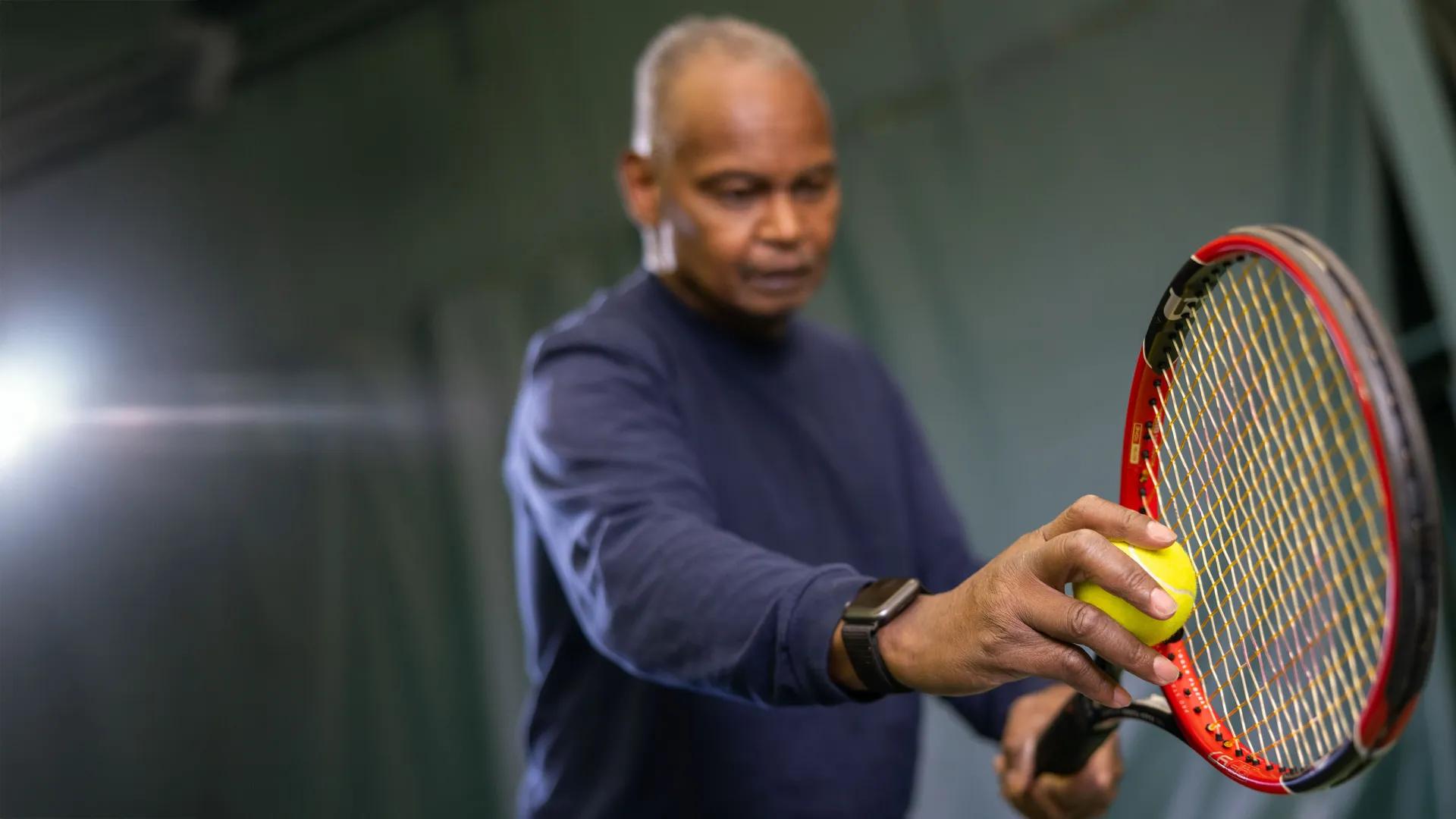 Older black man wearing navy blue shirt and holding a red tennis racket and yellow tennis ball with arms in front