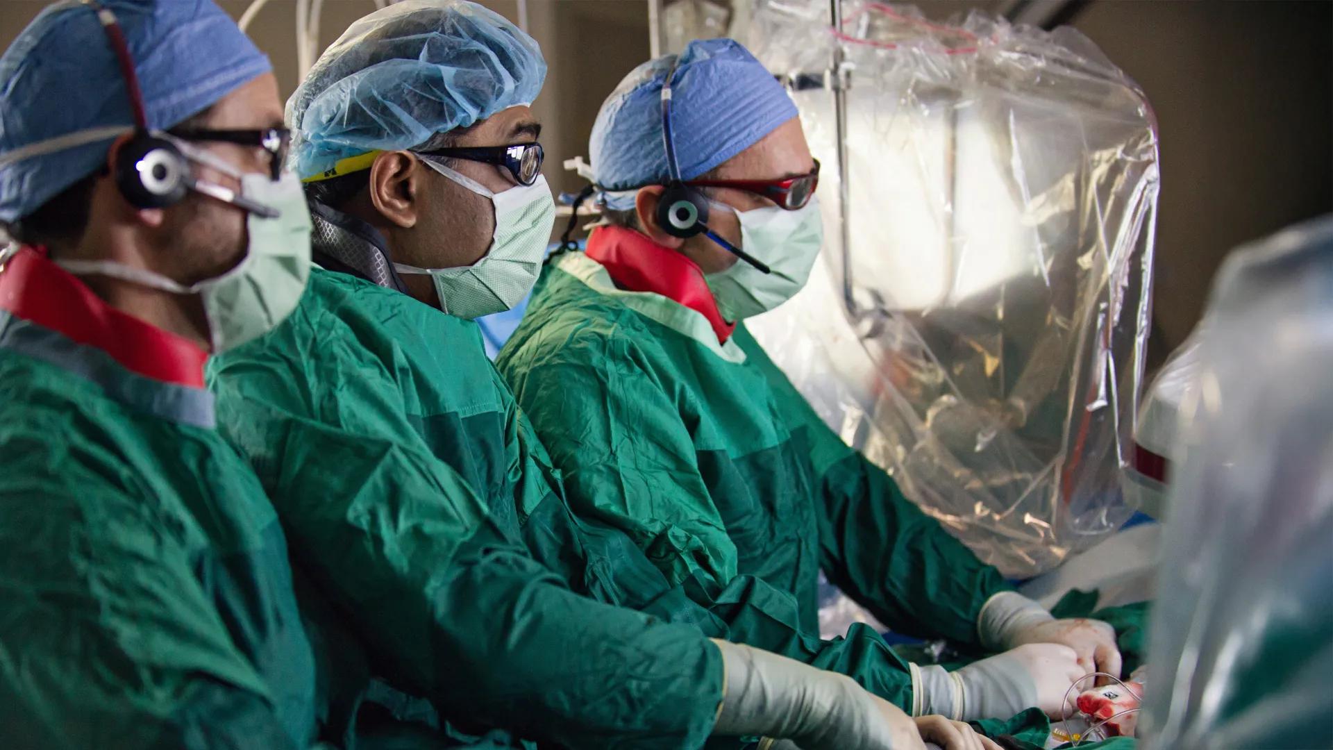 Three cardiac surgeons, gowned in green and masked, performing surgery