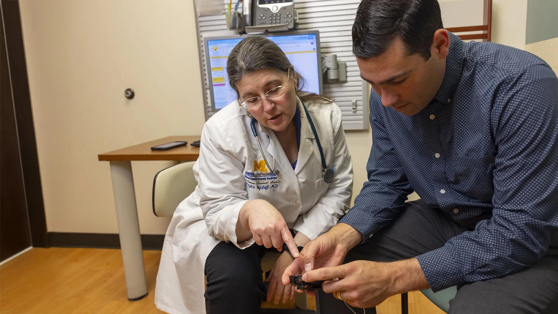 A doctor in a white coat with a stethoscope sits next to a patient in a clinic exam room, pointing at a glucose meter the patient is holding while explaining how to use it.