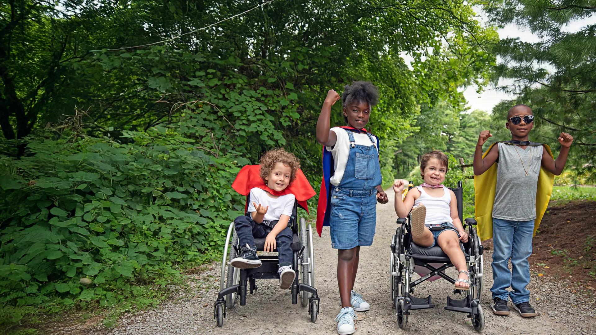 Dressed as superheroes, two wheelchair bound children smile and pose with other kids.