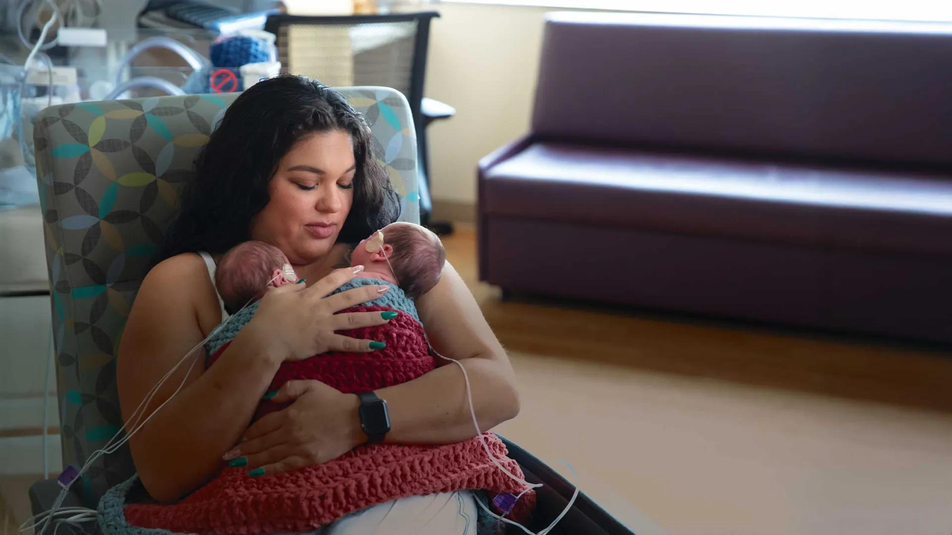 a mother sits in a chair holding her two premature infants